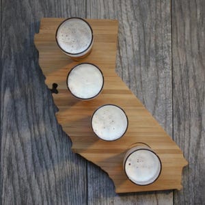 May include: A wooden beer flight board shaped like the state of California, holding four glasses of beer. The glasses are filled with a light-colored beer and topped with white foam. The board sits on a gray wooden surface.