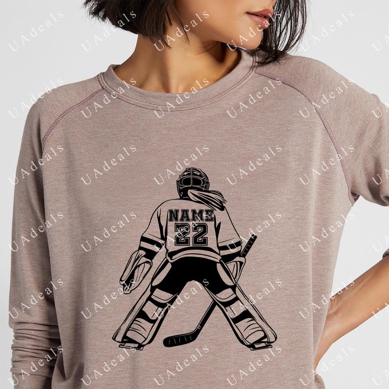 A digital graphic of a female hockey goalie seen from behind, wearing a jersey with the placeholder 'NAME' and the number '22'. The design is black on a soft, muted pink sweatshirt, showcasing a sporty and personalized style.