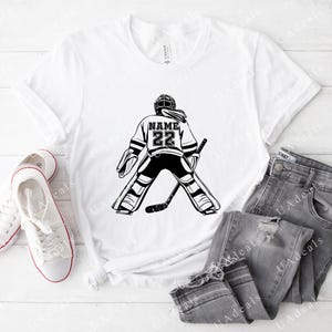 A white t-shirt featuring a black graphic of a female hockey goalie from the back, wearing a jersey with the number '22' and a placeholder for 'NAME.' The shirt is styled with gray jeans and white sneakers on a light wooden surface.