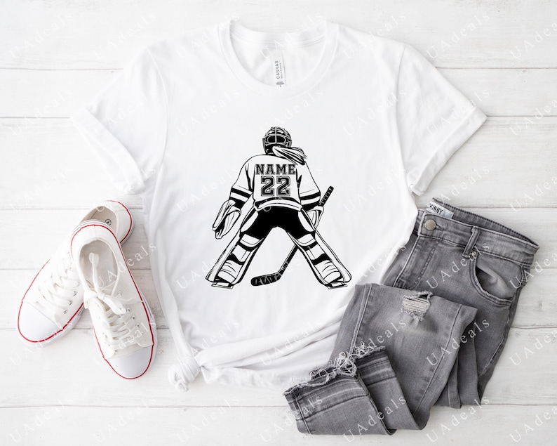A white t-shirt featuring a black graphic of a female hockey goalie from the back, wearing a jersey with the number '22' and a placeholder for 'NAME.' The shirt is styled with gray jeans and white sneakers on a light wooden surface.