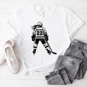 May include: White t-shirt featuring a black and white graphic of a hockey player wearing a jersey with the name "Olivia" and the number "22".