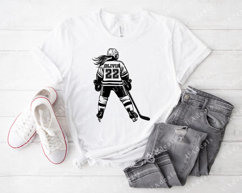 May include: White t-shirt featuring a black and white graphic of a hockey player wearing a jersey with the name "Olivia" and the number "22".