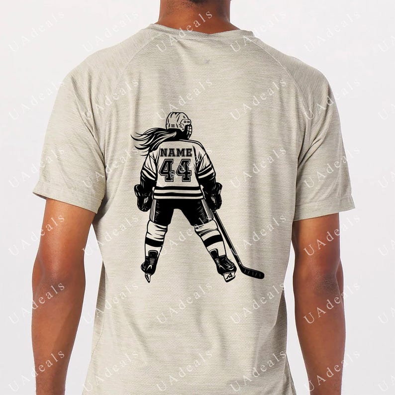 May include: A light grey t-shirt featuring a black and white graphic of a hockey player. The player is wearing a jersey with the number 44 and the word "NAME" above it. The player is holding a hockey stick and is in a skating position.