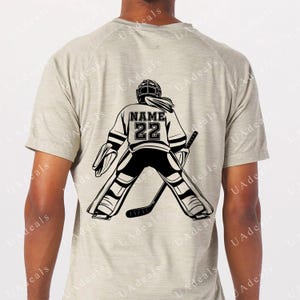A digital graphic of a female hockey goalie seen from behind, wearing a jersey with the placeholder 'NAME' and the number '22'. The design is in black on a light gray shirt, showcasing the goalie stance with a stick.