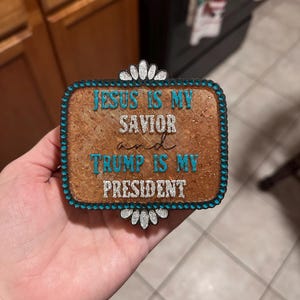 May include: A wooden sign with a turquoise border and silver glitter lettering. The sign reads "Jesus is my savior and Trump is my president."