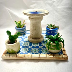 May include: A miniature ceramic bird bath with a white base and a blue bowl, surrounded by four small potted plants on a blue and white tiled tray with a wooden frame.