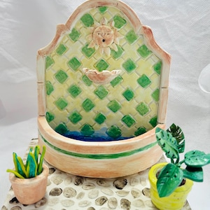 Puede incluir: Una fuente de cerámica en miniatura con una pared de azulejos amarillos y verdes, una cara de sol y una pequeña piscina. La fuente está sobre una base con un patrón de piedra y dos pequeñas plantas en maceta.