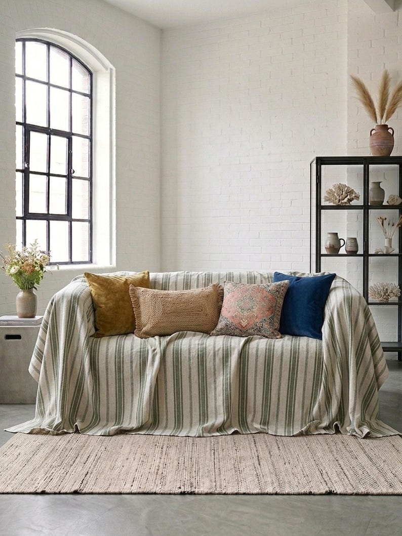 May include: A living room scene featuring a sofa draped with a striped throw and several decorative pillows in shades of gold, brown, pink, and blue. A woven rug and a black display cabinet complete the cozy, neutral-toned space.