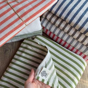 May include: A collection of folded striped kitchen towels in various colors, including red, blue, green, and gray. The towels are neatly stacked, with one bundle tied with twine. The towels have a classic, farmhouse style.