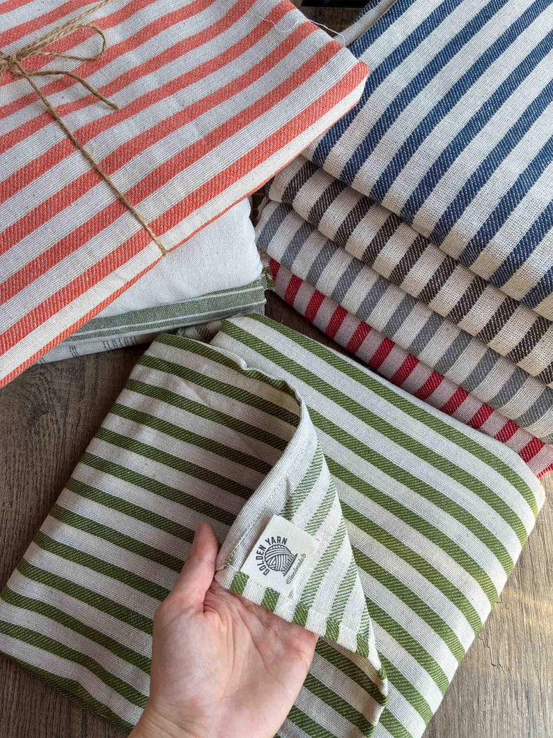 May include: A collection of folded striped kitchen towels in various colors, including red, blue, green, and gray. The towels are neatly stacked, with one bundle tied with twine. The towels have a classic, farmhouse style.