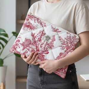 May include: A quilted zippered pouch featuring a white fabric base with a red toile de jouy pattern depicting pastoral scenes. A small rectangular label with the text Golden Yarn is stitched onto the center of the front panel.