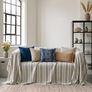 May include: A sofa with a striped throw and several decorative pillows in shades of blue, beige, and tan. The pillows have various textures and patterns. A woven rug is on the floor in front of the sofa. A large window is in the background.