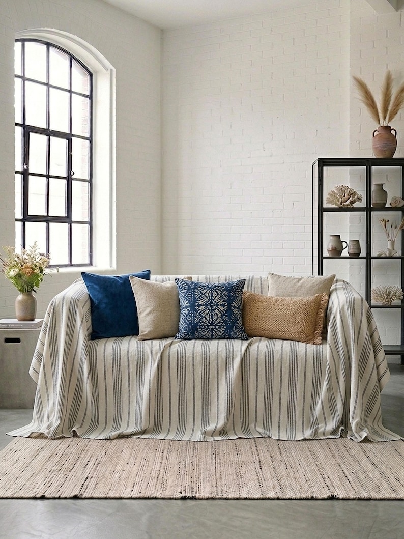 May include: A sofa with a striped throw and several decorative pillows in shades of blue, beige, and tan. The pillows have various textures and patterns. A woven rug is on the floor in front of the sofa. A large window is in the background.
