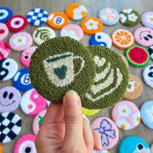 May include: Hand-tufted, round coasters with various designs. One pair features a green background with a coffee cup and whipped cream design. Other coasters have designs like an 8-ball, smiley face, and a yin-yang symbol. The coasters are made of a textured, looped material.