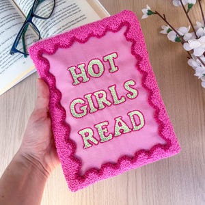 May include: A pink book cover with the text "HOT GIRLS READ" in green lettering. The cover has a textured, scalloped border. A pair of glasses and an open book are in the background.