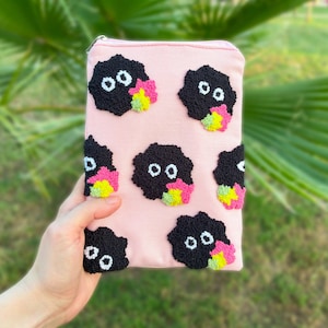 Op de afbeelding: Een roze etui met ritssluiting met geborduurde zwarte roetgeesten met witte ogen en kleurrijke accenten. De etui wordt in een hand gehouden tegen een vage groene achtergrond, met een grillig ontwerp.
