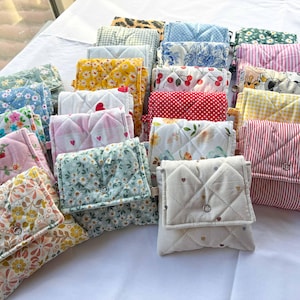 May include: A collection of small, quilted fabric pouches featuring various patterns including floral prints, gingham, polka dots, stripes, cherries, and animal motifs. Each rectangular pouch has a fold-over flap secured with a single metal snap button.
