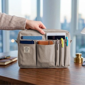 Könnte beinhalten: Eine beige Organizer-Tasche mit mehreren Taschen, die ein Tablet, Stifte und Notizbücher enthält. Die Tasche hat ein Etikett mit der Aufschrift "GOLDEN YARN". Die Tasche befindet sich auf einer Holzoberfläche, mit einem Notizbuch und einem Stift im Hintergrund.