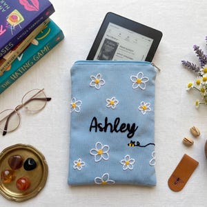 Könnte beinhalten: Eine hellblaue E-Reader-Hülle mit weißen Gänseblümchen und dem Namen "Ashley". Die Hülle enthält einen schwarzen E-Reader. Weitere Artikel sind Bücher, Brille, eine kleine Schale mit Steinen, ein Leder-Lesezeichen und eine Schüssel mit Pistazien.