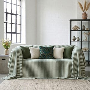 Puede incluir: Un salón con un sofá cubierto por una manta a rayas verdes y blancas. El sofá tiene varios cojines en tonos verdes, crema y turquesa. Una alfombra tejida y una vitrina negra completan la escena.