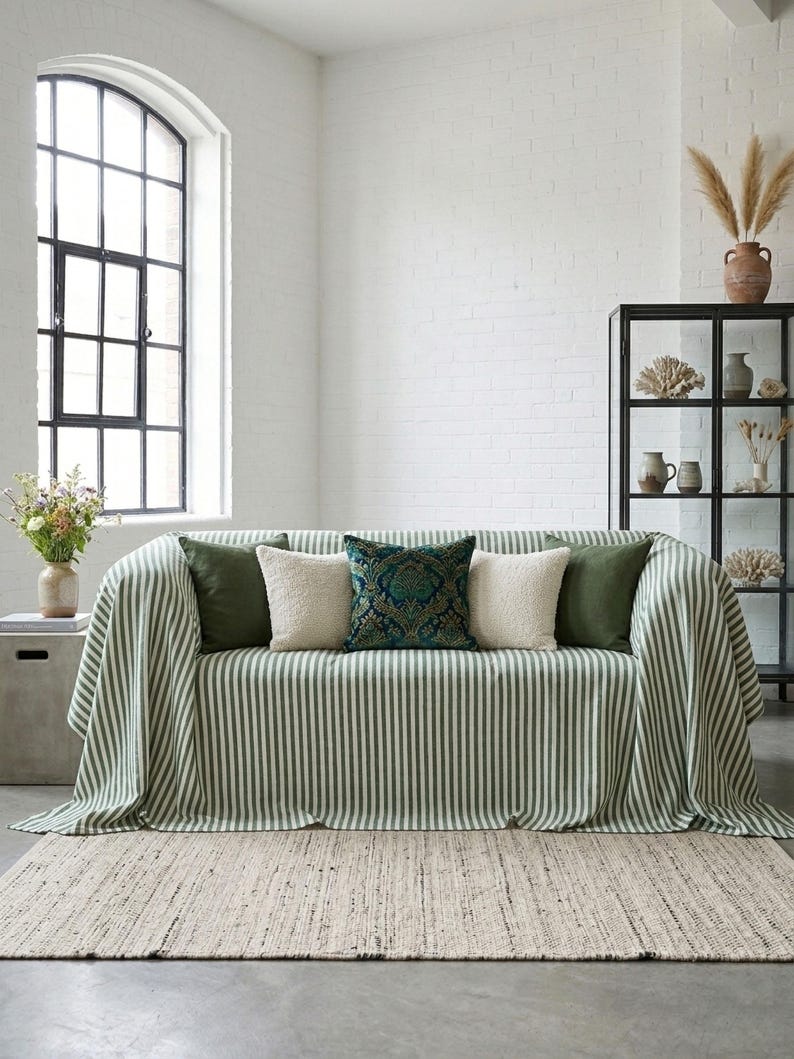 May include: A living room scene featuring a sofa draped with a green and white striped throw. The sofa is adorned with several decorative pillows in shades of green, cream, and teal. A woven rug and a black display cabinet complete the cozy interior.