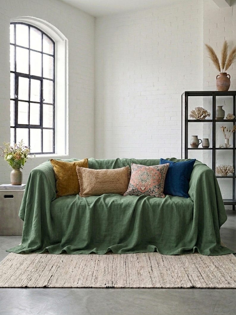 May include: A green sofa with several decorative pillows in shades of gold, tan, pink, and blue. The sofa is covered with a green throw, and sits on a natural fiber rug. A black metal shelf and large window are in the background.