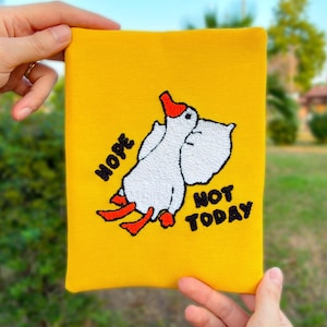 May include: Yellow pouch with an embroidered design of a white goose with a red beak and feet, and the words "Nope Not Today" in black. The pouch is held by hands.