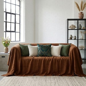 Könnte beinhalten: Ein braunes Sofa, das mit einer passenden Decke bedeckt ist. Das Sofa ist mit dekorativen Kissen in Grün-, Weiß- und Türkistönen ausgestattet. Ein gewebter Teppich liegt vor dem Sofa. Im Hintergrund sind ein großes Fenster und ein schwarzes Regal zu sehen.