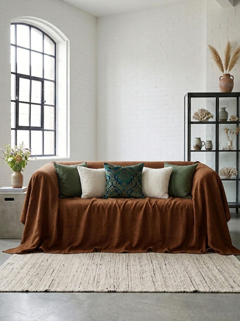 May include: A brown sofa covered with a matching throw blanket. The sofa is adorned with five decorative pillows in shades of green, white, and teal. A woven rug sits on the floor in front of the sofa. The room has a large window and a black shelving unit.