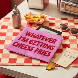 May include: A light purple zippered pouch featuring the red embroidered text "WHATEVER I'M GETTING CHEESE FRIES" on the front. The pouch is rectangular with a silver zipper closure and is positioned on a red and white checkered surface.