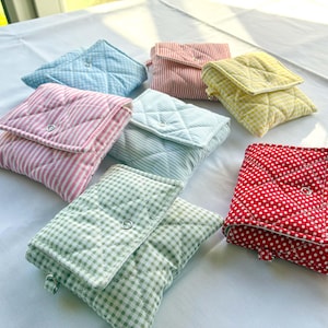 May include: A collection of seven small, quilted fabric pouches featuring various patterns including gingham, stripes, and polka dots in colors such as red, yellow, light blue, pink, and green. Each square pouch has a flap secured with a metal snap.