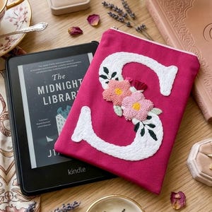 Może przedstawiać: Różowa kosmetyczka zapinana na zamek z dużą, białą, haftowaną literą „S” i kwiatowymi akcentami. Czytnik Kindle wyświetlający „The Midnight Library” spoczywa obok kosmetyczki. Inne przedmioty to naszyjnik, filiżanka do herbaty i dekoracyjna miseczka.