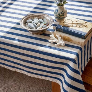 May include: A blue and white striped tablecloth covers a wooden table. A silver bowl filled with smooth stones sits on the tablecloth, along with a small vase of flowers, two books, and a pair of round glasses. A piece of white coral is also on the table.