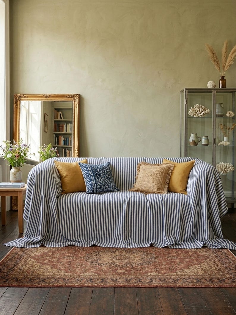 May include: A living room scene featuring a sofa draped with a black and white striped throw. The sofa is adorned with several decorative pillows in shades of gold and blue. A patterned rug lies on the wooden floor, and a gold-framed mirror hangs on the wall.