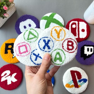 May include: A collection of colorful, round coasters with gaming-related designs. The coasters feature symbols from PlayStation, Xbox, Nintendo Switch, and Twitch, along with controller button icons. The coasters are white with colorful embroidered details.