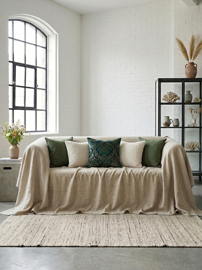 May include: A beige sofa with a matching throw blanket and several decorative pillows in shades of green and cream. The sofa sits on a neutral-toned rug in a bright, airy room with a large window and a black display cabinet.
