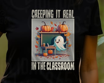 May include: A black t-shirt with a white graphic of a ghost sitting at a desk with pumpkins and autumn leaves. The text on the shirt reads "CREEPING IT REAL IN THE CLASSROOM".