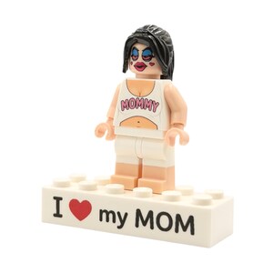 Puede incluir: Una figura de Lego de una mujer con cabello negro, que lleva una camiseta blanca con "MOMMY" impresa y pantalones cortos blancos. La figura se encuentra sobre una base de ladrillo Lego blanco con "I ❤️ my MOM" impreso. La figura tiene maquillaje.