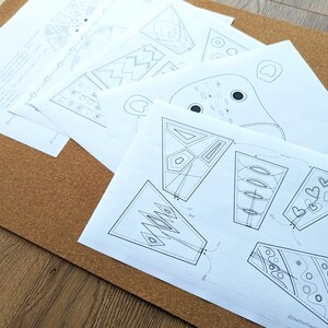 May include: A collection of white paper sheets with black line drawings of geometric shapes and animal faces. The papers are arranged on a brown corkboard surface, with a wooden table visible in the background. The drawings appear to be patterns or templates.