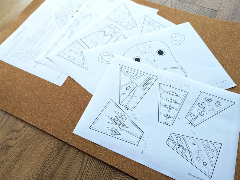 May include: A collection of white paper sheets with black line drawings of geometric shapes and animal faces. The papers are arranged on a brown corkboard surface, with a wooden table visible in the background. The drawings appear to be patterns or templates.