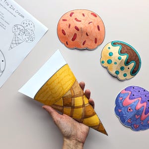 Op de afbeelding: Een hand houdt een papieren ijshoorntje vast naast drie kleurrijke papieren ijsbollen. De bollen zijn oranje, bruin en paars met decoratieve details. Er ligt ook een vel papier met illustraties en instructies voor ijshoorntjes.
