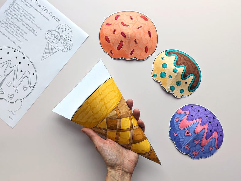 Op de afbeelding: Een hand houdt een papieren ijshoorntje vast naast drie kleurrijke papieren ijsbollen. De bollen zijn oranje, bruin en paars met decoratieve details. Er ligt ook een vel papier met illustraties en instructies voor ijshoorntjes.