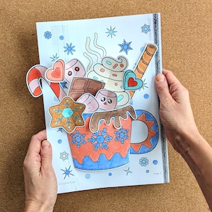 Peut inclure: Illustration color&eacute;e d'une tasse de chocolat chaud avec des guimauves, une canne en sucre et un biscuit pain d'&eacute;pices. La tasse est d&eacute;cor&eacute;e de flocons de neige et d'une anse bleue. L'image est tenue par deux mains.
