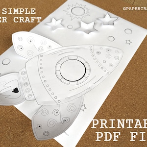 May include: Printable 3D paper craft of a rocket ship with a large window, a flame, and a star. The rocket is surrounded by stars and a sun. The text "Printable PDF File" is at the bottom of the page.