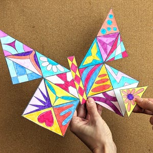 May include: A colorful, geometric butterfly design with various shapes and patterns. The wings feature triangles, hearts, and floral elements in shades of pink, blue, yellow, and purple. The butterfly is held by a person's hands.