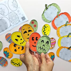 Op de afbeelding: Een verzameling papieren knutselwerkjes met diverse ovale vormen met kleurrijke patronen en pompoengezichten, naast verschillende pompoenvormige sjablonen en een instructieblad met de titel Pumpkins Craft en een QR-code.