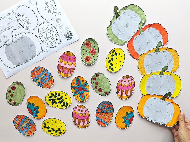 Op de afbeelding: Een papieren knutselset met gedrukte sjablonen voor pompoenen en ovale vormen. De set bevat een wit instructieblad met zwarte lijntekeningen en meerdere kleurrijke, met de hand uitgeknipte papieren ovalen en pompoenen met diverse patronen.