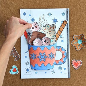 Peut inclure: Illustration color&eacute;e d'une tasse de chocolat chaud avec des guimauves, de la cr&egrave;me fouett&eacute;e et une gaufrette. La tasse est d&eacute;cor&eacute;e de flocons de neige et d'un motif bleu et orange. Formes d&eacute;coup&eacute;es suppl&eacute;mentaires : &eacute;toile en pain d'&eacute;pices et c&oelig;ur.