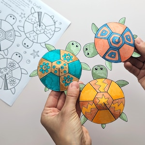 Op de afbeelding: Kleurrijk papieren schildpaddenknutselwerk met oranje, blauwe en gele schilden, vastgehouden door handen. De schildpadden hebben groene vinnen en koppen. Op de achtergrond een blad met schildpadcontouren, getiteld "Turtles Craft".