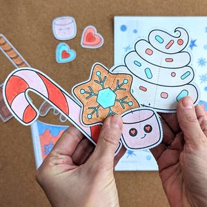 Peut inclure: Ensemble de d&eacute;coupes de papier sur le th&egrave;me de No&euml;l, comprenant une canne en bonbon, une &eacute;toile en pain d'&eacute;pices, une guimauve et une spirale de cr&egrave;me fouett&eacute;e avec des vermicelles. Les d&eacute;coupes sont tenues devant un livret partiellement visible.